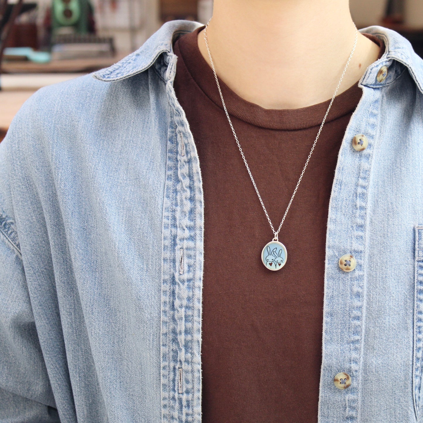 A silver pendant necklace with a circular charm with a rabbit design, worn by a person over a brown t-shirt and a light blue denim shirt.