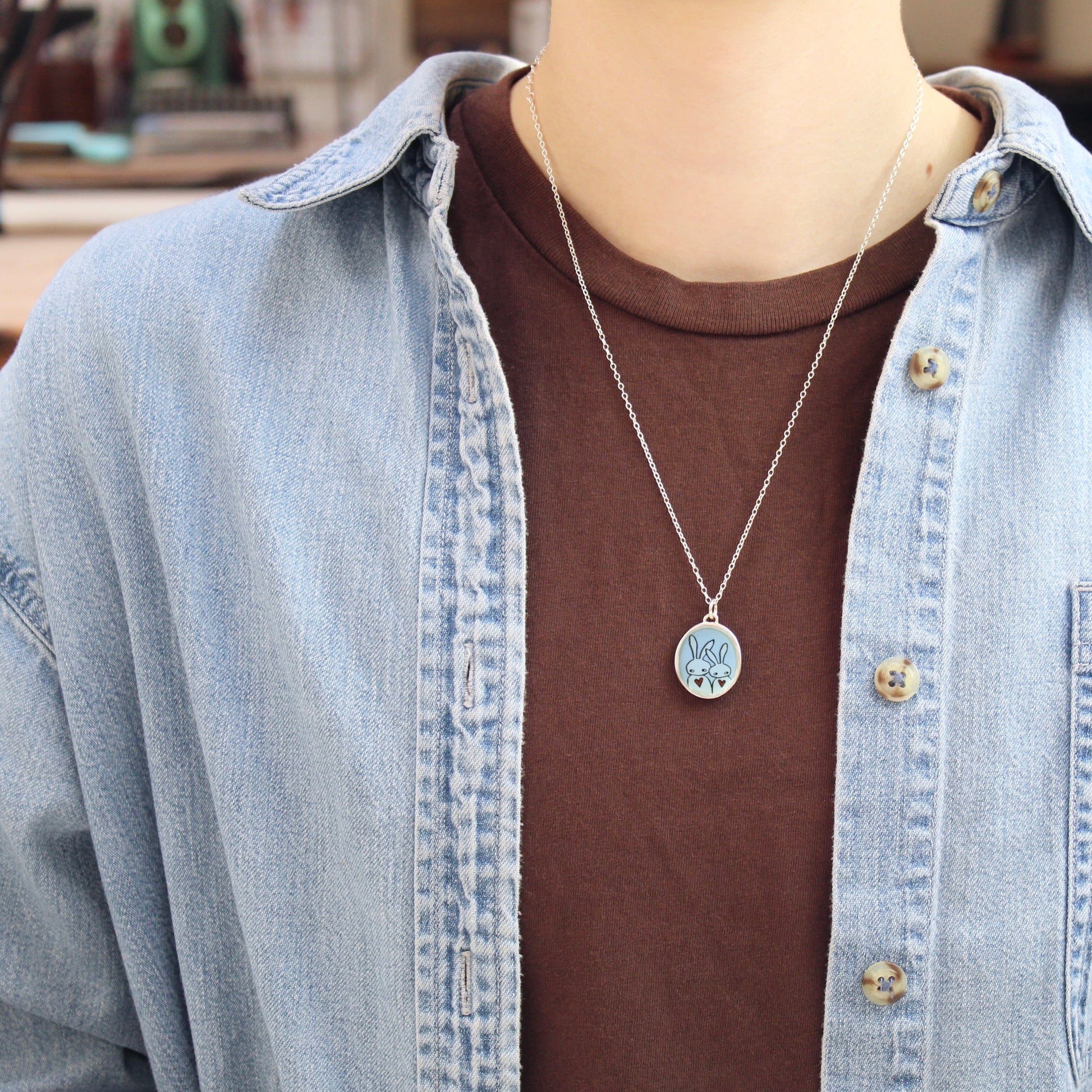 A silver pendant necklace with a circular charm with a rabbit design, worn by a person over a brown t-shirt and a light blue denim shirt.