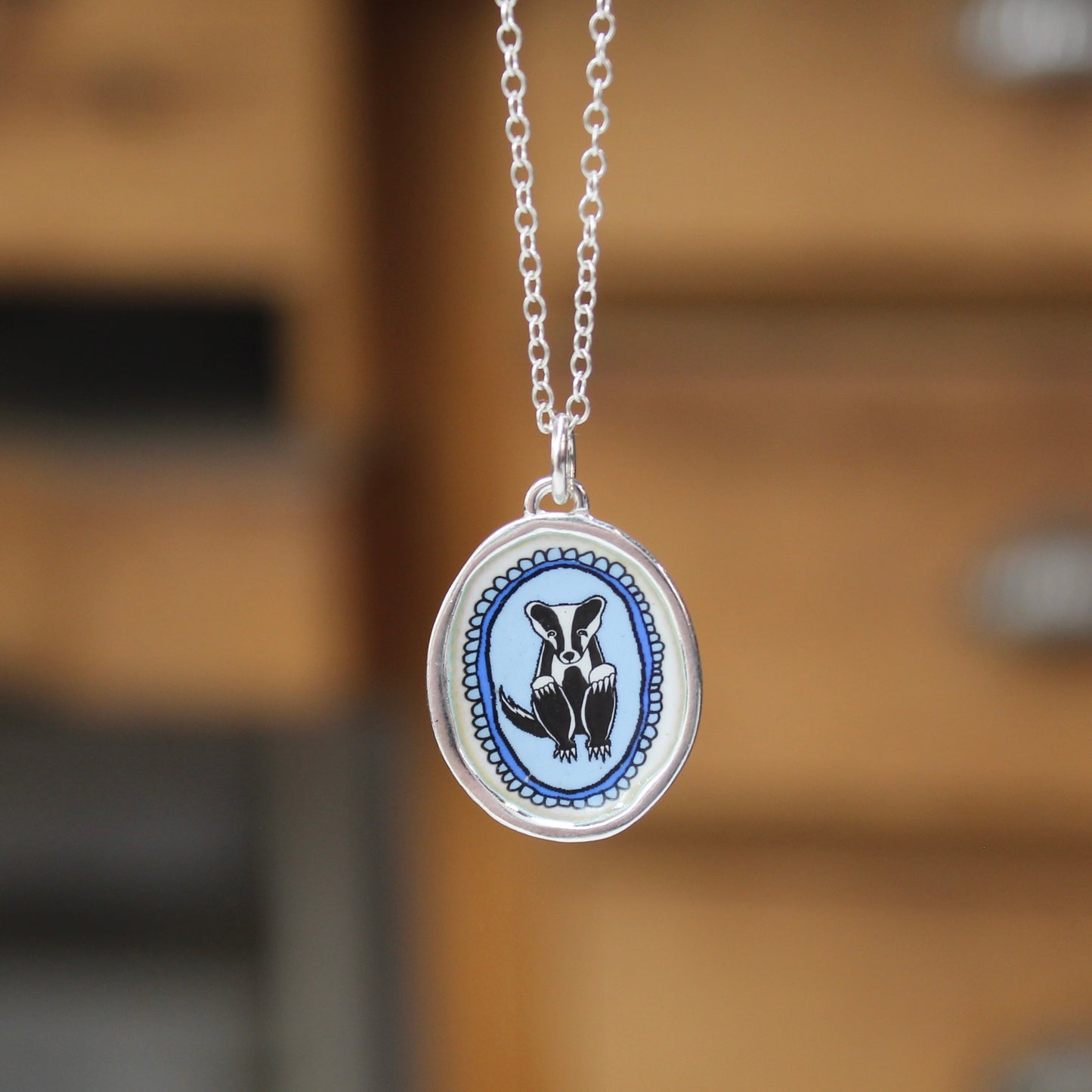 A silver oval pendant necklace featuring a hand-painted badger illustration on a light blue background, suspended from a silver chain against a blurred wooden background.