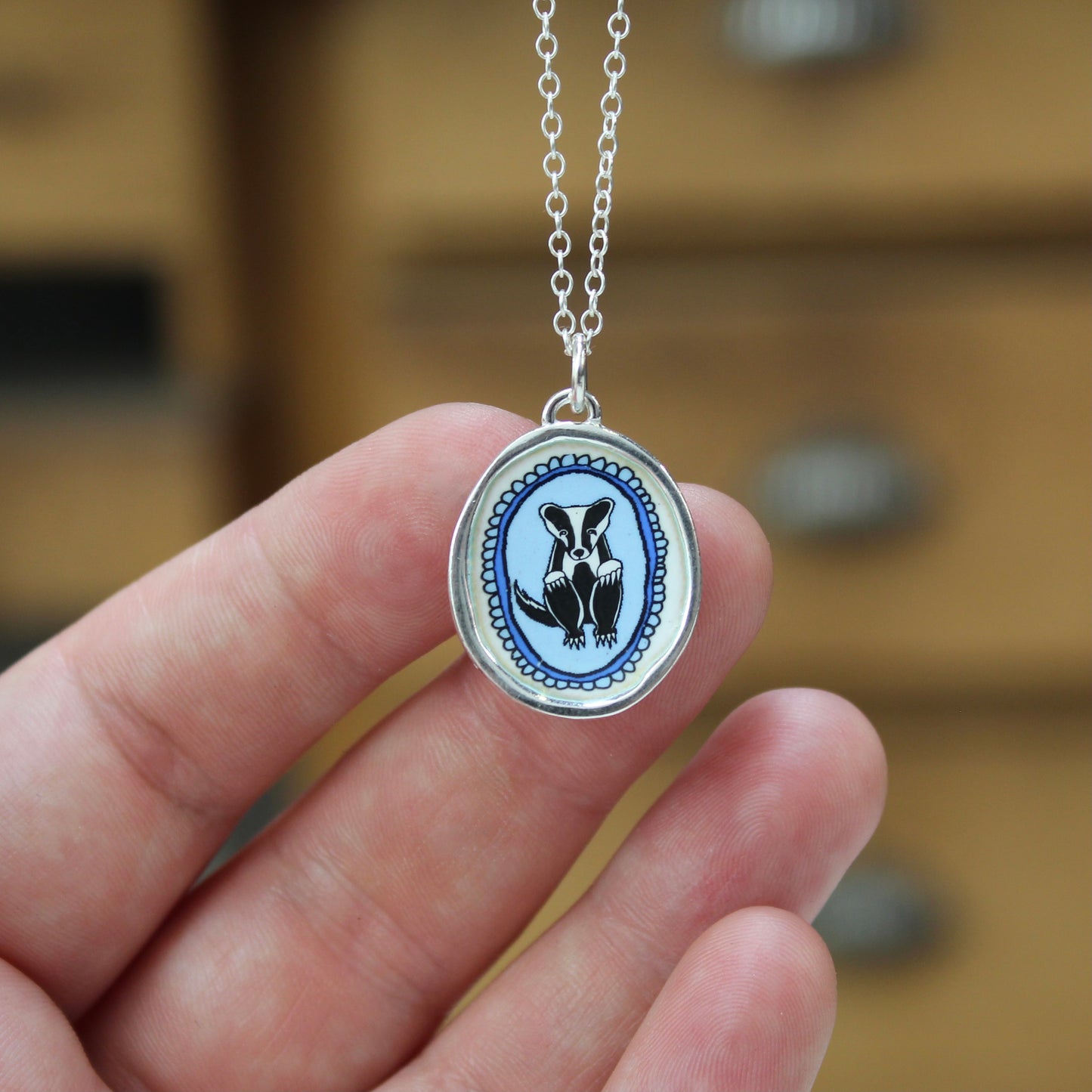 A hand holding a silver oval pendant necklace featuring a blue and black badger design, with a silver chain against a blurred wooden background.