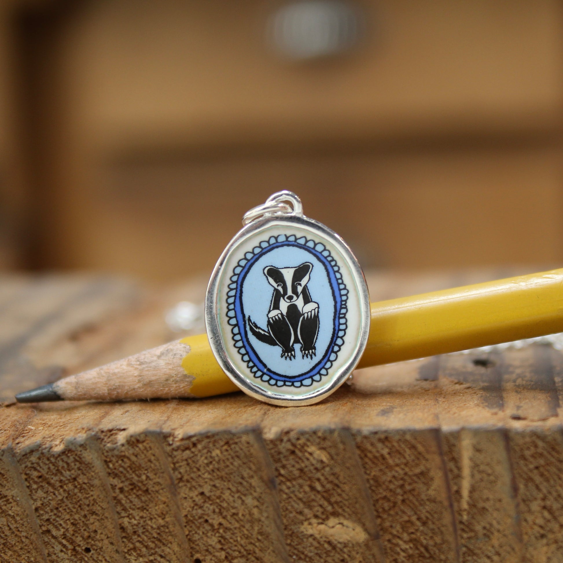 An oval silver pendant featuring a black and white badger illustration on a light blue background, resting on a wooden surface next to a yellow pencil.