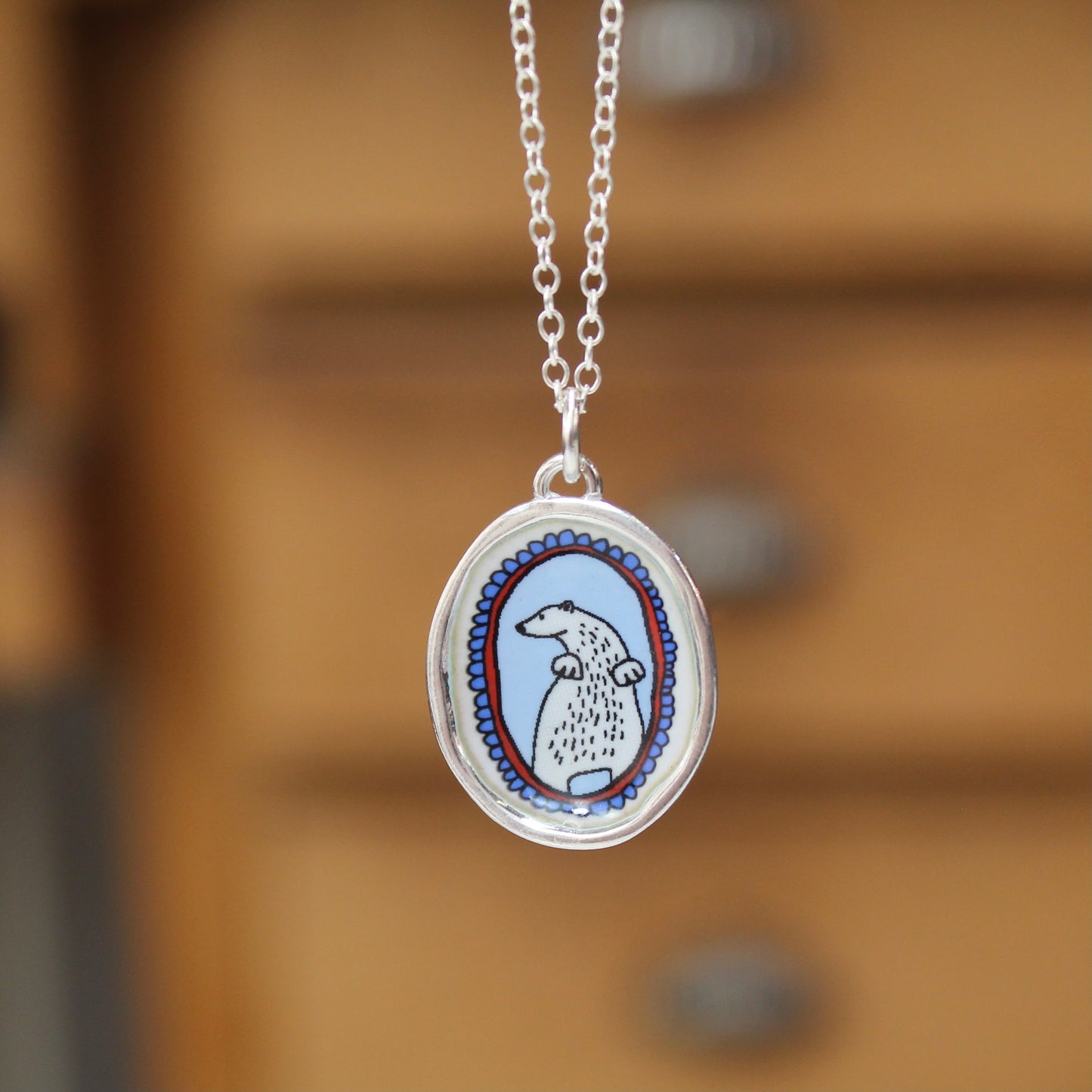 A silver oval pendant necklace featuring a hand-drawn style illustration of a white dog with black details, set against a light blue background with red and blue decorative borders, hanging against a blurred warm brown wooden background.