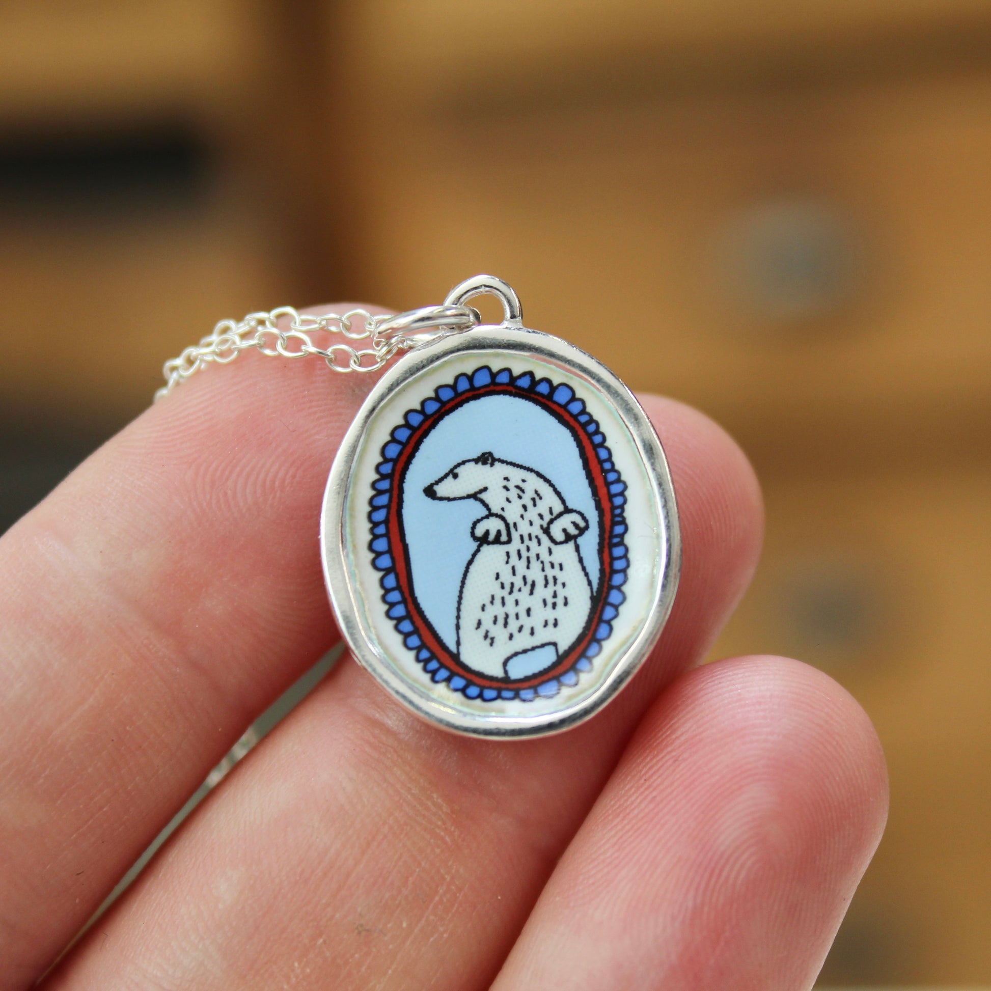A close-up of a hand holding a silver oval pendant necklace featuring a stylized white polar bear on a light blue background with a blue and red patterned border.