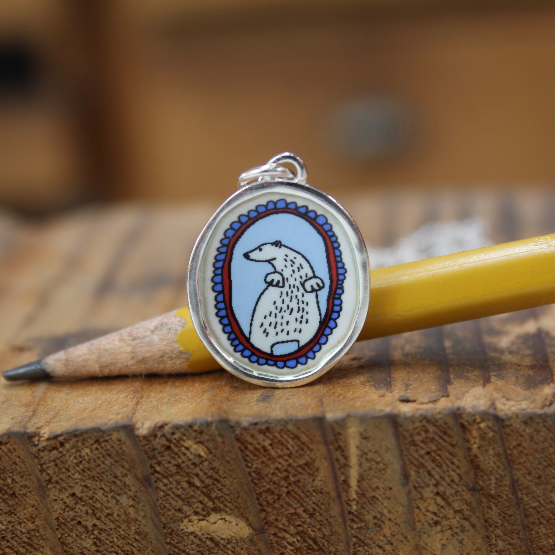 An oval pendant charm featuring a cartoon polar bear illustration, resting on a wooden surface next to a yellow pencil.