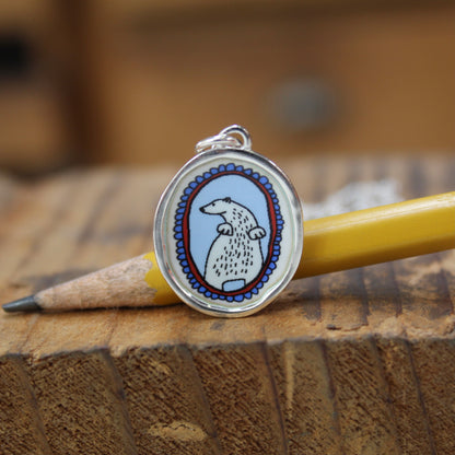 An oval pendant charm featuring a cartoon polar bear illustration, resting on a wooden surface next to a yellow pencil.