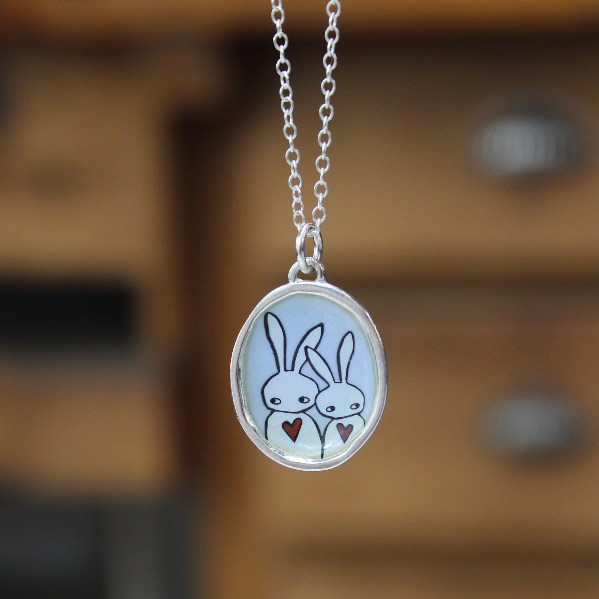 A silver oval pendant necklace featuring a cartoon illustration of two white rabbits holding small red hearts, hanging against a blurred warm brown wooden background.