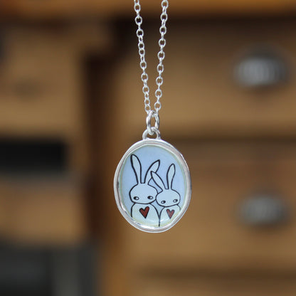 A silver oval pendant necklace featuring a cartoon illustration of two white rabbits holding small red hearts, hanging against a blurred warm brown wooden background.