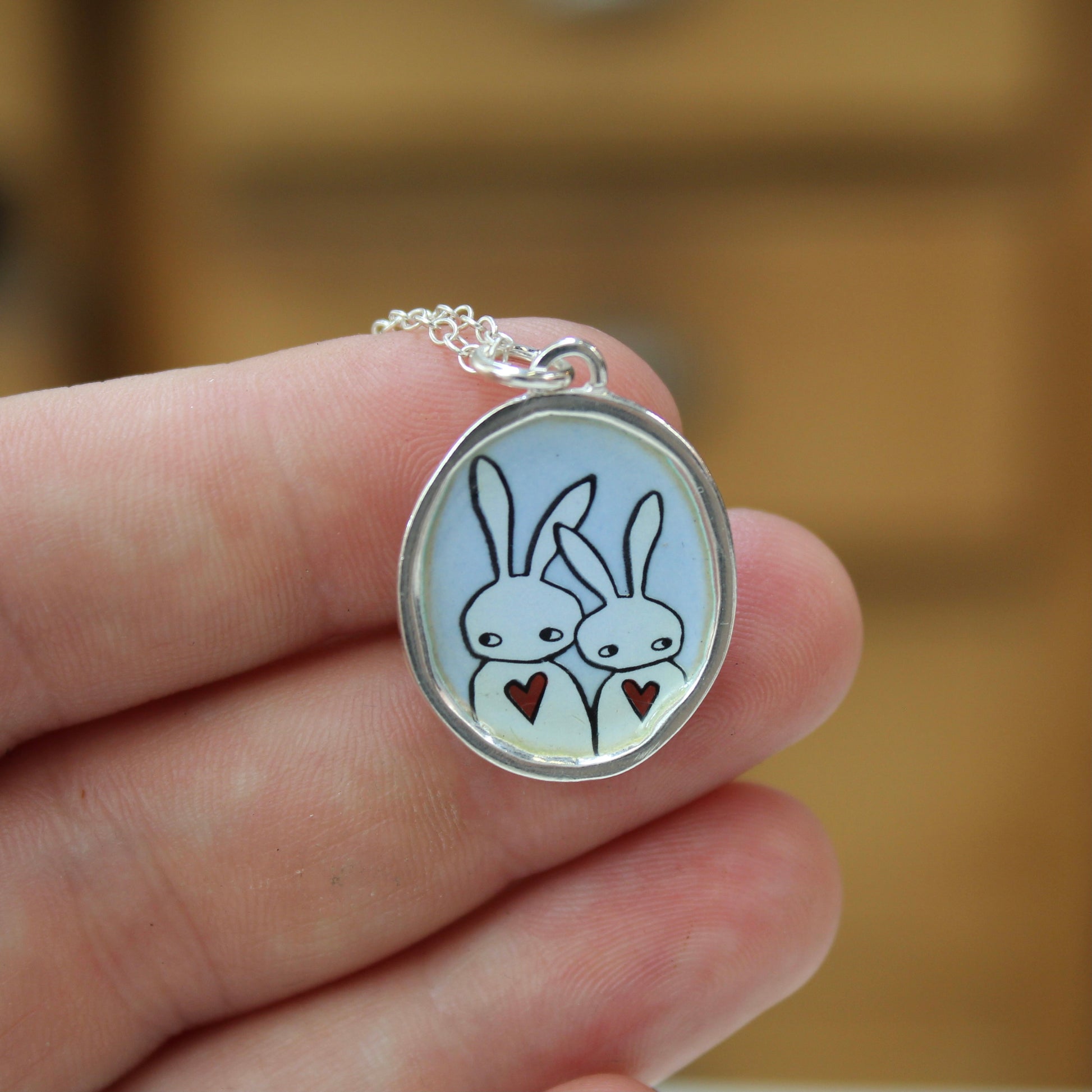 A silver oval pendant necklace featuring a cartoon bunny illustration, held in a person's hand against a blurred wooden background.
