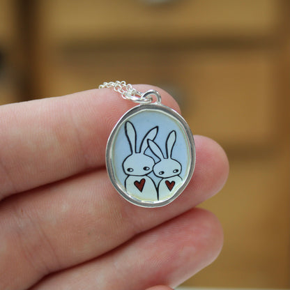 A silver oval pendant necklace featuring a cartoon bunny illustration, held in a person's hand against a blurred wooden background.
