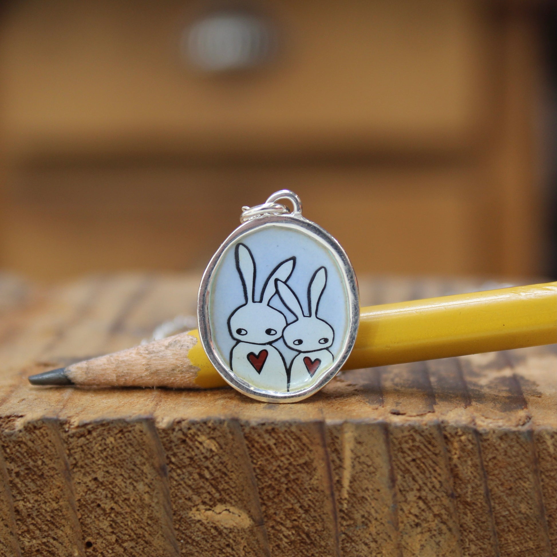 An oval pendant charm with a silver border, featuring two white cartoon rabbits holding red hearts on a pale blue background, resting on a yellow pencil atop a wooden block.