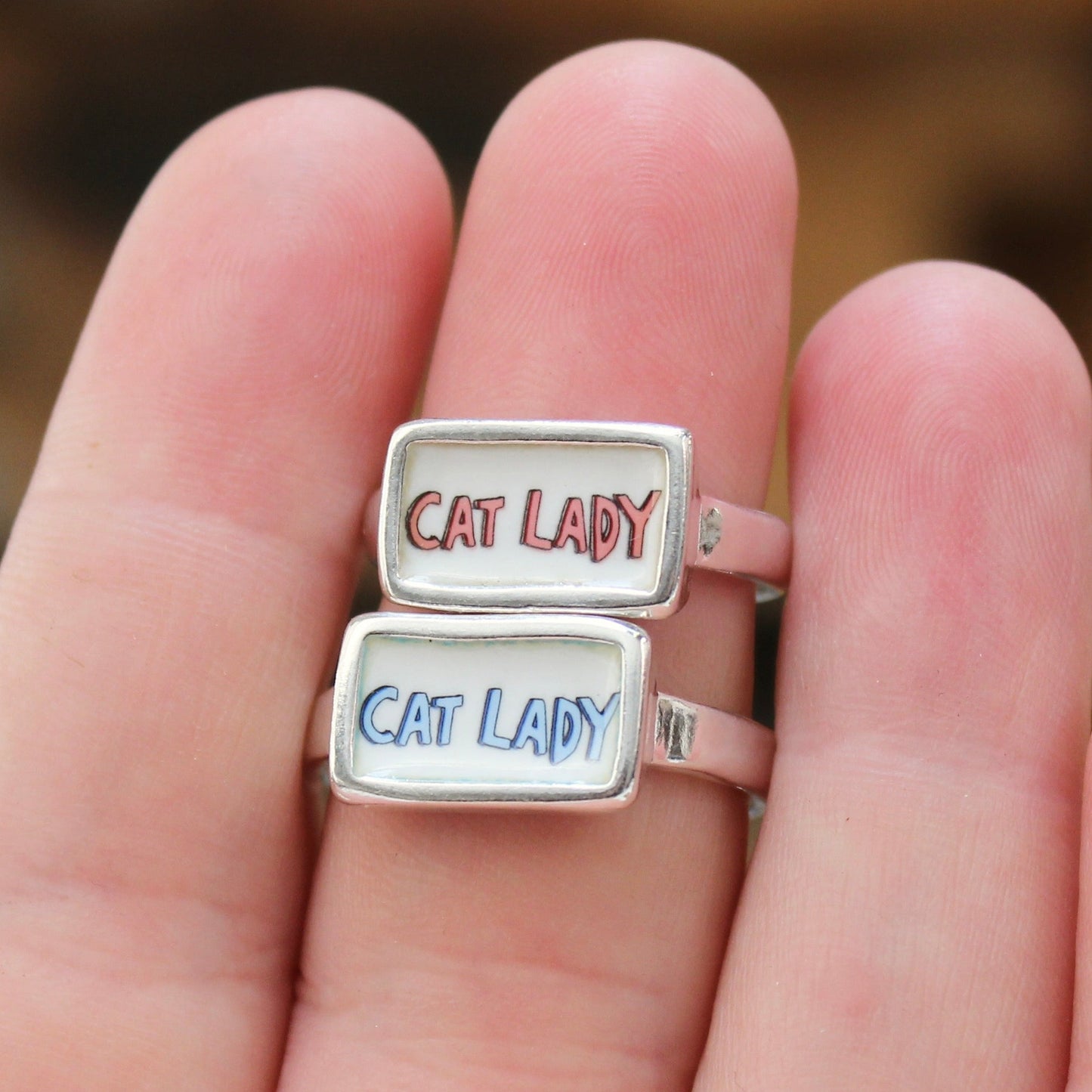 Two silver rings with white rectangular faces, one with 'CAT LADY' in red and the other in blue, worn on a person's fingers.