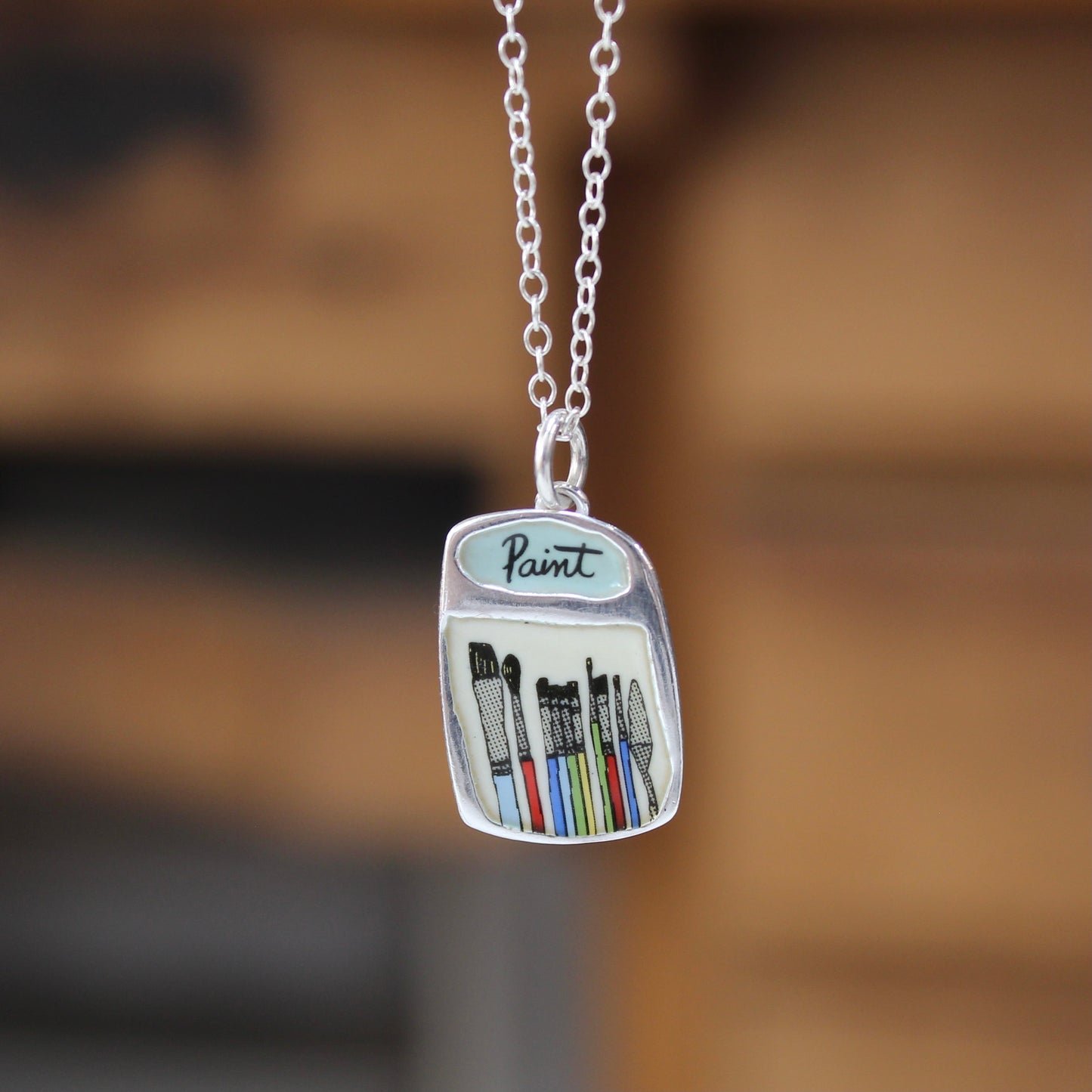 A silver pendant necklace shaped like a paint can, featuring the word 'Paint' and colorful illustrated paintbrushes, hanging against a blurred warm brown background.