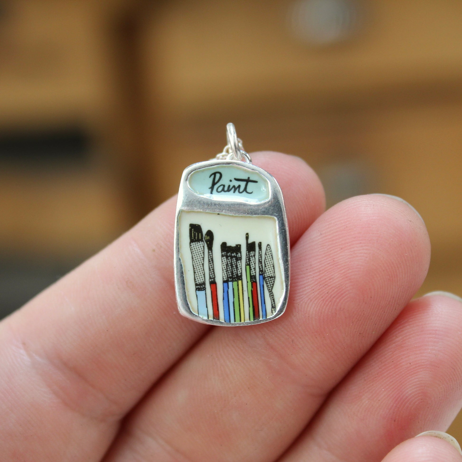 A hand holding a small silver pendant shaped like a paint palette, with the word 'Paint' on top and illustrated paintbrushes in multiple colors inside.