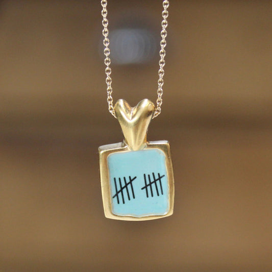 A gold pendant necklace featuring a square light blue enamel charm with black tally marks, suspended from a delicate gold chain against a warm, blurred brown background.
