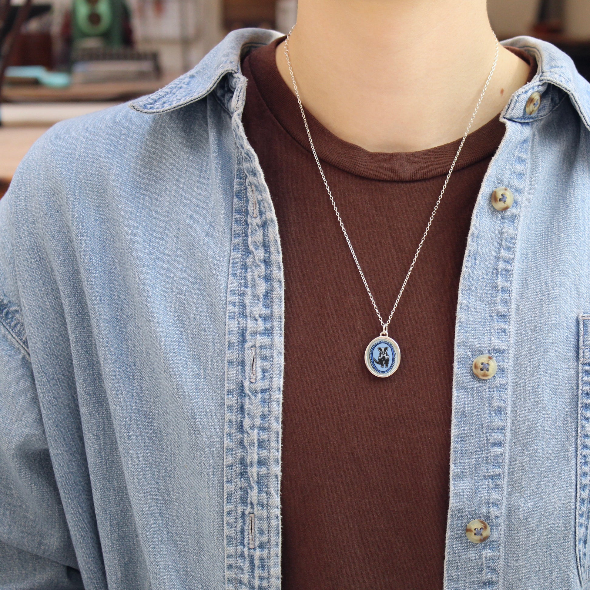 A silver oval pendant necklace with a blue and white badger design, worn on a person's neck over a brown t-shirt and a light blue denim shirt.