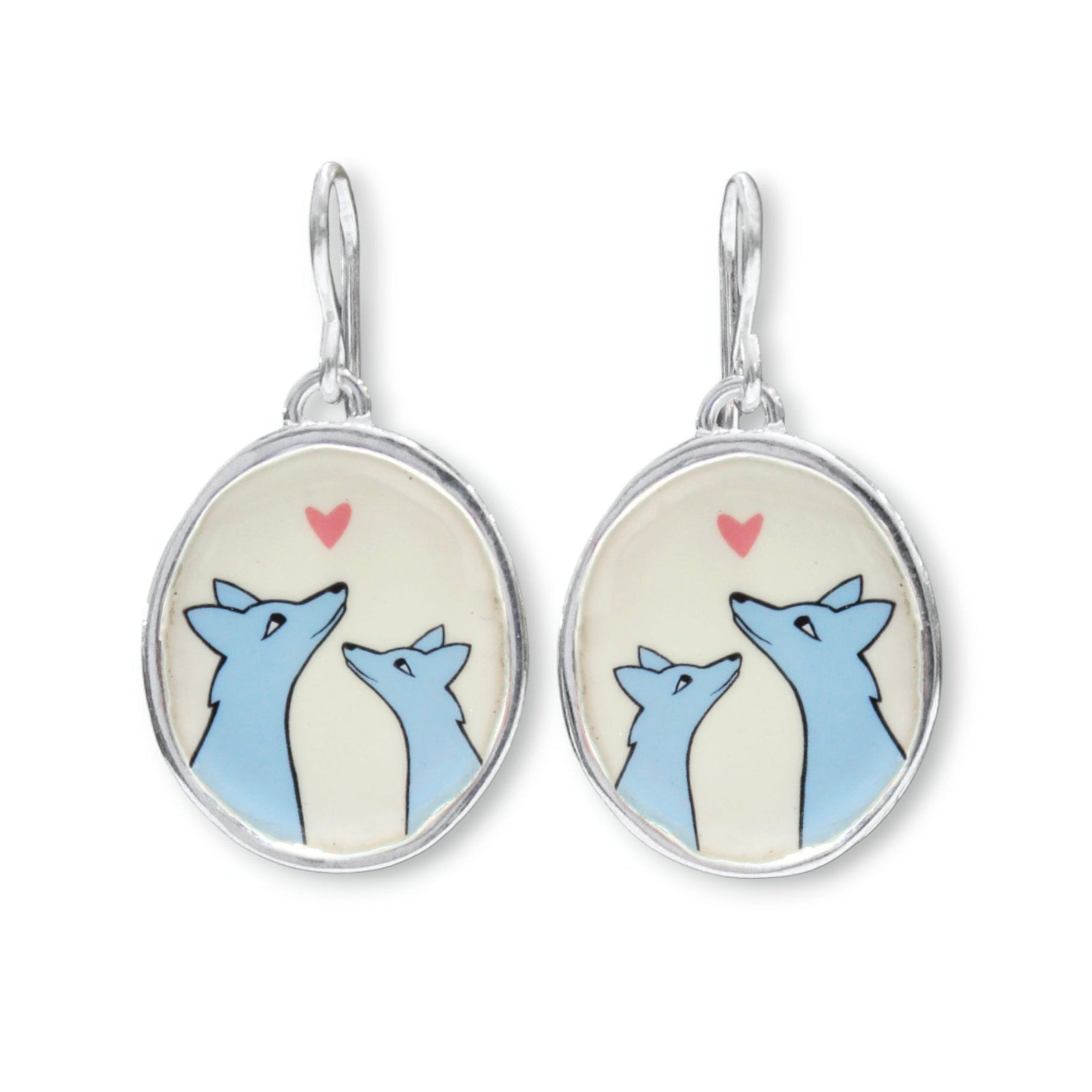 A pair of silver dangle earrings with oval charms featuring a cartoon illustration of two blue foxes looking at each other under a small pink heart on a cream background.