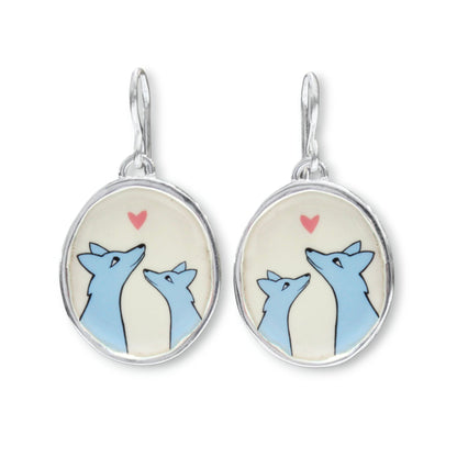 A pair of silver dangle earrings with oval charms featuring a cartoon illustration of two blue foxes looking at each other under a small pink heart on a cream background.