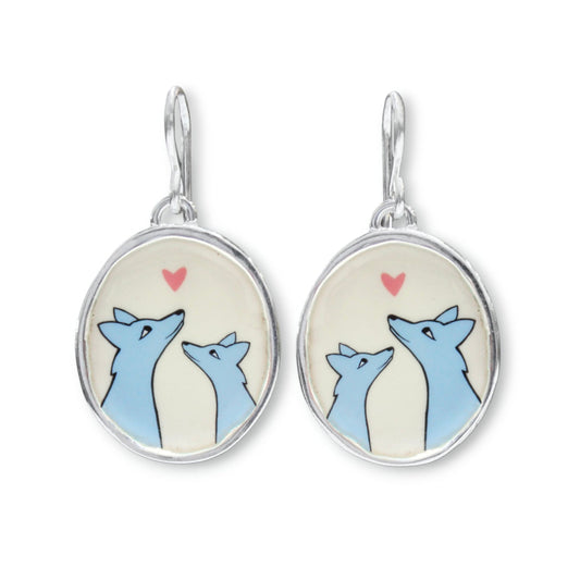 A pair of silver dangle earrings with oval charms featuring a cartoon illustration of two blue foxes looking at each other under a small pink heart on a cream background.