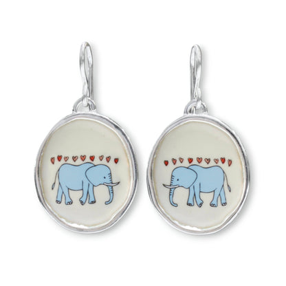 A pair of silver drop earrings featuring oval charms with a light blue elephant and small red hearts on a cream background.