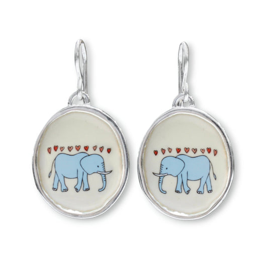 A pair of silver drop earrings featuring oval charms with a light blue elephant and small red hearts on a cream background.