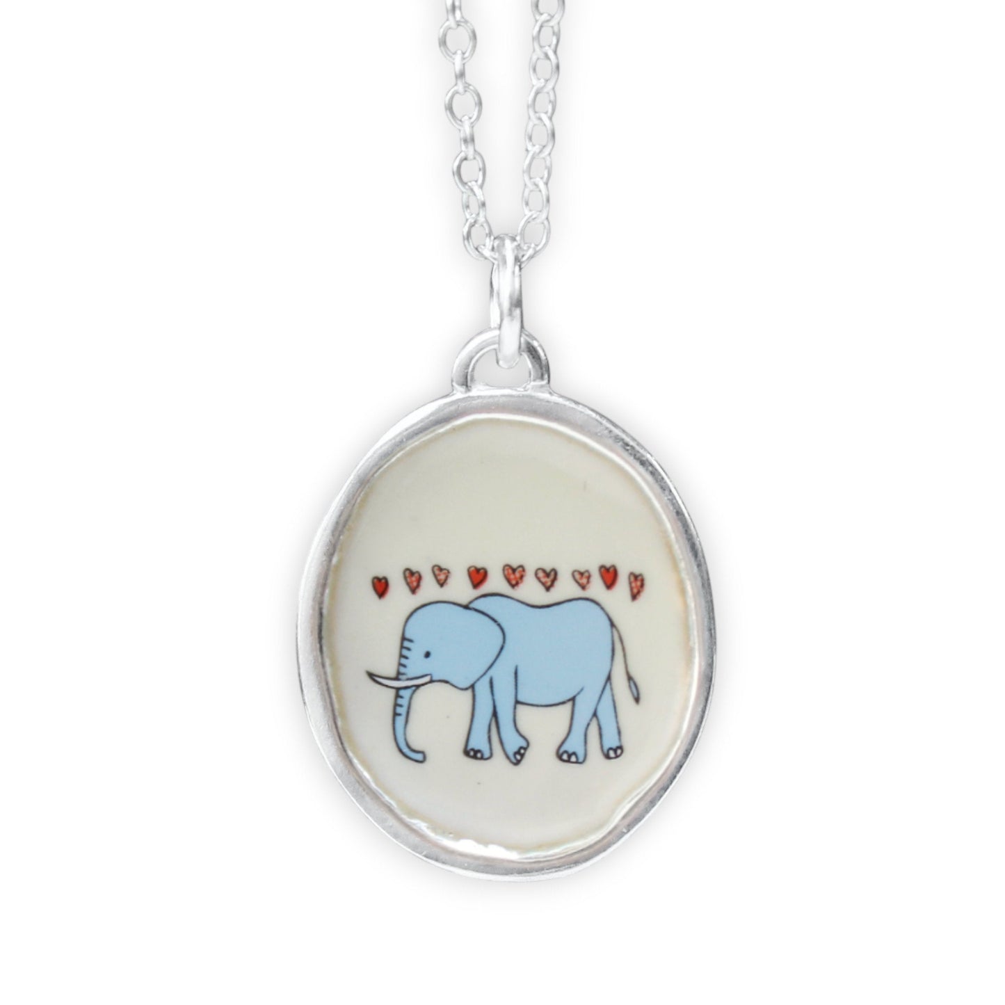 A silver oval pendant necklace featuring a light blue cartoon elephant under small red hearts on a cream background, shown against a plain white studio background.
