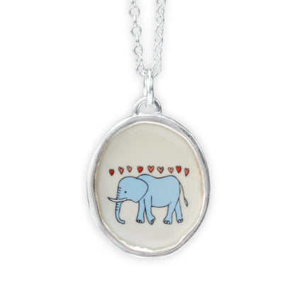 A silver oval pendant necklace featuring a light blue cartoon elephant under small red hearts on a cream background, shown against a plain white studio background.