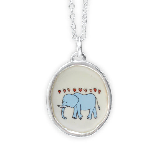 A silver oval pendant necklace featuring a light blue cartoon elephant under small red hearts on a cream background, shown against a plain white studio background.
