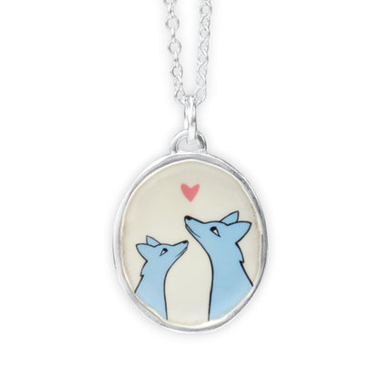 A silver oval pendant necklace featuring a cute illustration of two blue foxes looking up at a small pink heart on a cream background, suspended from a silver chain against a white background.