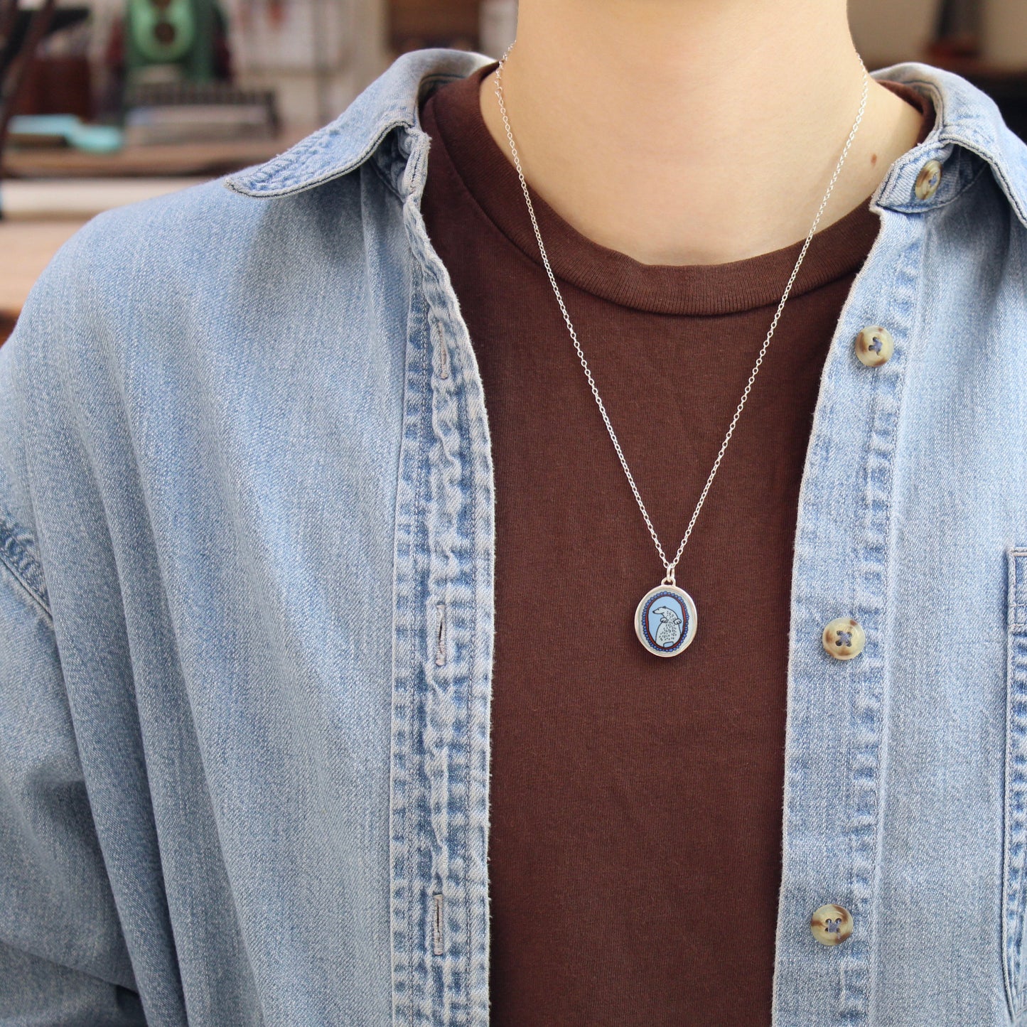 A silver oval pendant necklace with a blue and white polar bear design, worn on a person's neck over a brown t-shirt and a light blue denim shirt.