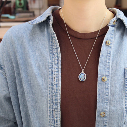 A silver oval pendant necklace with a blue and white polar bear design, worn on a person's neck over a brown t-shirt and a light blue denim shirt.