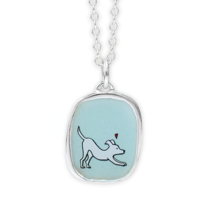 A silver pendant necklace featuring a light blue oval bezel with a white dog illustration and a small red heart, suspended from a silver chain on a white background.