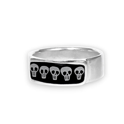 A silver-toned metal ring featuring a rectangular black inlay with five small white skull motifs, shown on a plain white background.