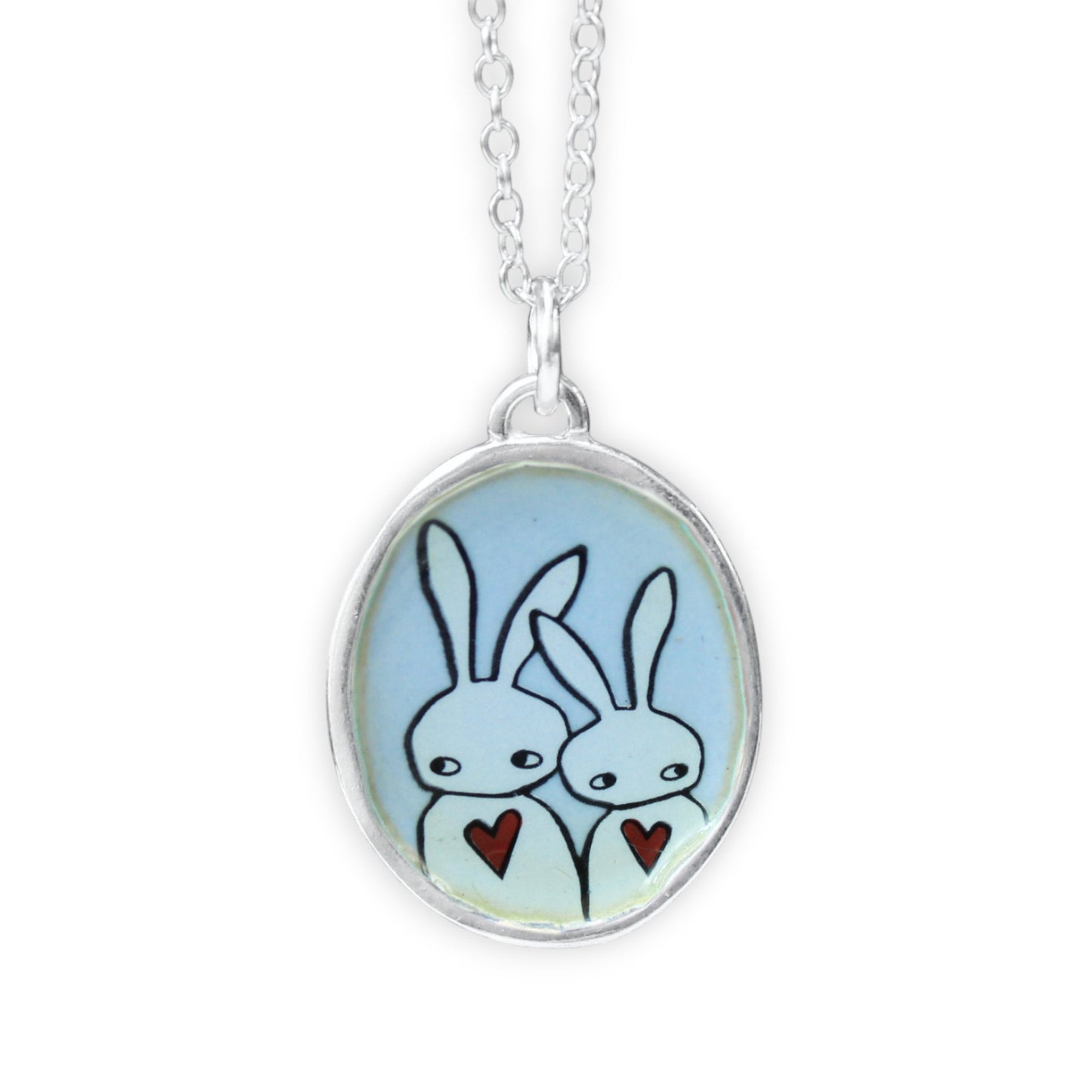 A silver oval pendant necklace featuring a light blue background with a hand-drawn illustration of two white rabbits holding small red hearts, suspended from a silver chain against a white background.