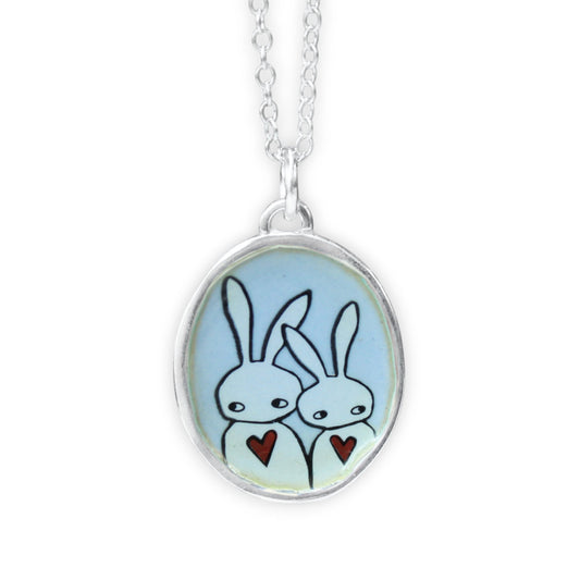 A silver oval pendant necklace featuring a light blue background with a hand-drawn illustration of two white rabbits holding small red hearts, suspended from a silver chain against a white background.