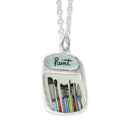 A silver pendant necklace shaped like a paint jar, featuring a light blue enamel top with the word 'Paint' and colorful illustrated paintbrushes inside, shown on a white background.