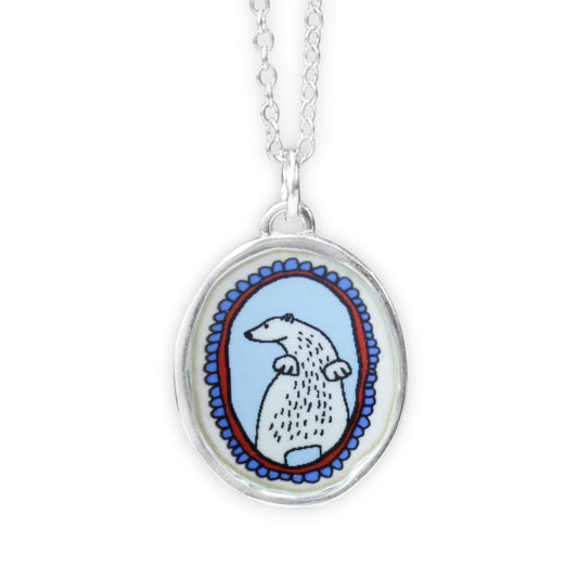 A silver oval pendant necklace featuring a hand-drawn style illustration of a white polar bear with black details, set against a light blue background with a red and blue patterned border, suspended from a silver chain on a white background.