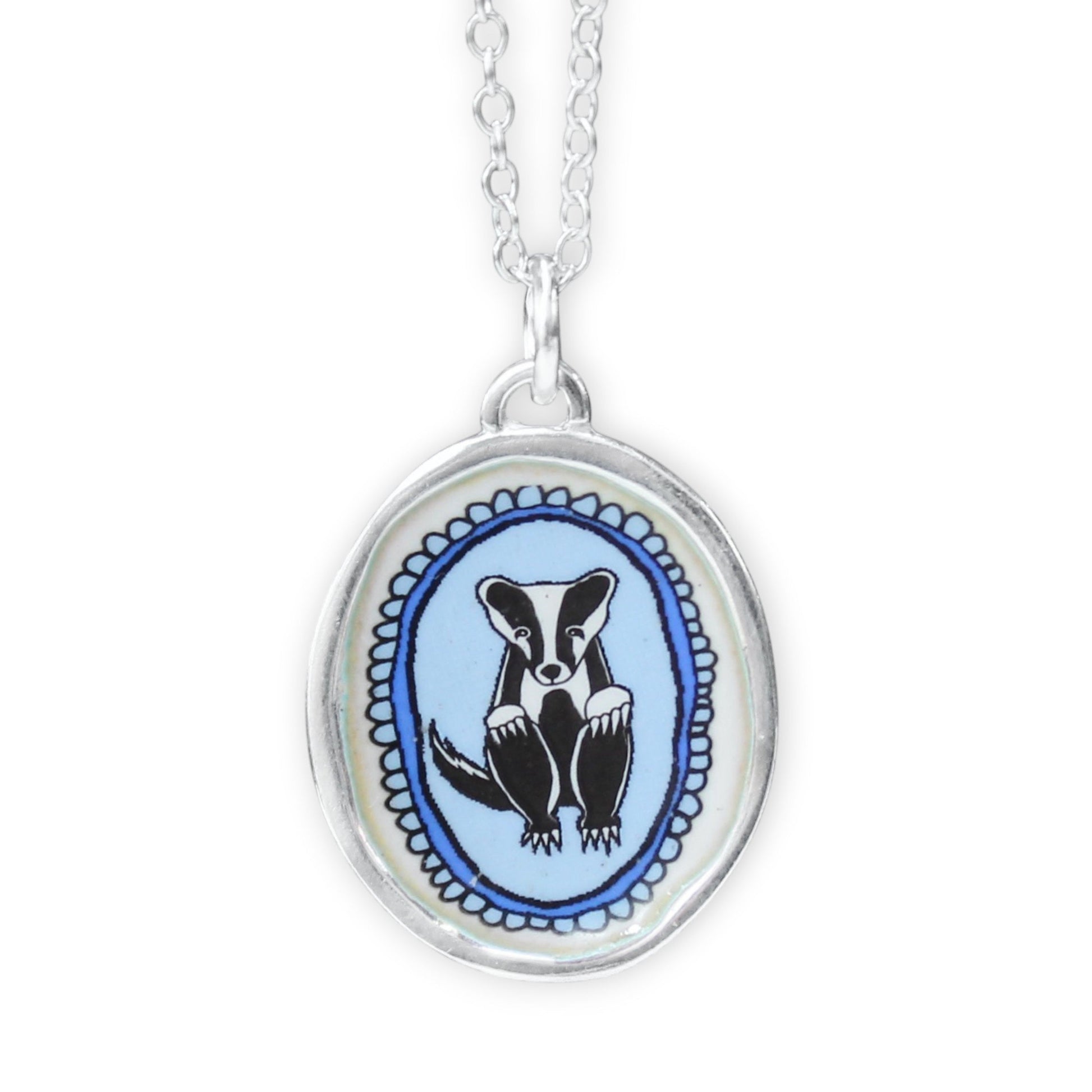 A silver oval pendant necklace featuring a black and white badger illustration on a light blue background, suspended from a silver chain against a white background.