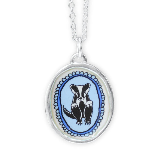 A silver oval pendant necklace featuring a black and white badger illustration on a light blue background, suspended from a silver chain against a white background.