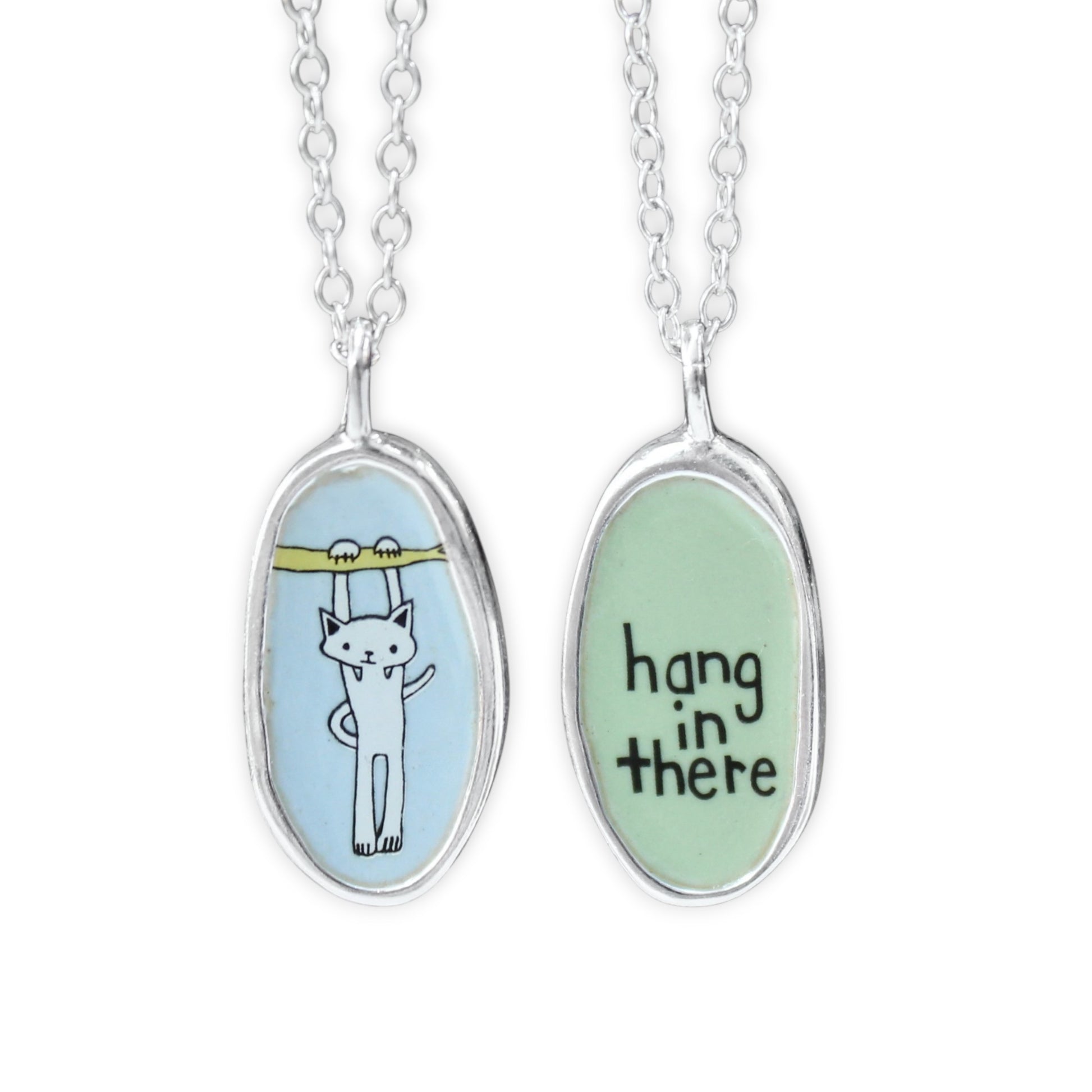Two silver oval pendant necklaces on a white background, showing the front and back of the same design. One pendant has a white cat illustration under a yellow bar on a light blue background, and the other has the words 'hang in there' in black script on a light green background.