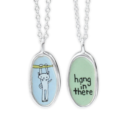 Two silver oval pendant necklaces on a white background, showing the front and back of the same design. One pendant has a white cat illustration under a yellow bar on a light blue background, and the other has the words 'hang in there' in black script on a light green background.